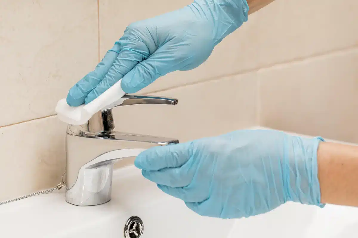 Gloved hands wiping a sink faucet with a cloth